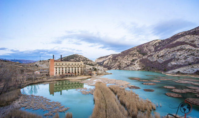 Panorama der Central Térmica de Aliaga am Embalse de Aliaga, das Kraftwerk spiegelt sich im Wasser zwischen kahlen Berghängen.