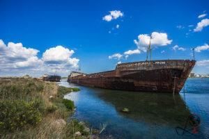 Verlassenes rostiges Frachtschiff am Ufer eines Hafenbeckens unter blauem Himmel mit weißen Wolken