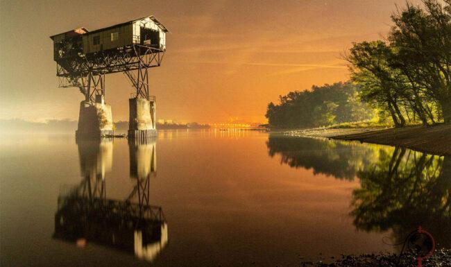 Der beleuchtete Kohleverladeplatz ragt nachts über dem Wasser auf; im Vordergrund liegt ein steiniger Donauuferstreifen.