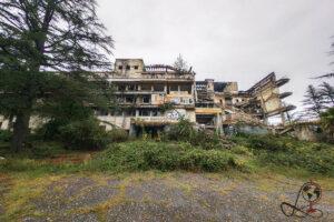 Das beschädigte Intourist-Hotel in Tskaltubo hinter Büschen und Bäumen, mit offenem Dachbereich und zerstörter Fassade.