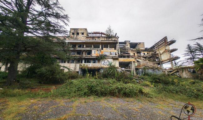 Das beschädigte Intourist-Hotel in Tskaltubo hinter Büschen und Bäumen, mit offenem Dachbereich und zerstörter Fassade.
