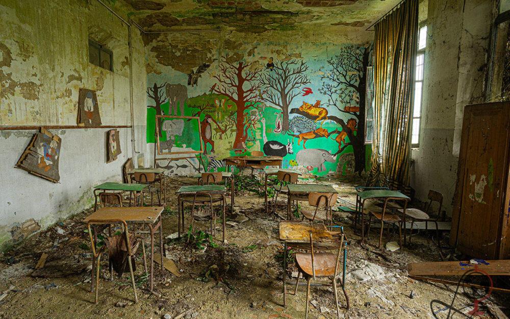 Klassenzimmer mit buntem Dschungelwandbild, rostigen Tischen und Stühlen, schwerem Vorhang und Pflanzen zwischen dem Schutt