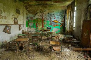 Klassenzimmer mit buntem Dschungelwandbild, rostigen Tischen und Stühlen, schwerem Vorhang und Pflanzen zwischen dem Schutt