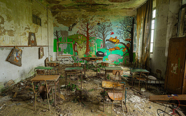 Klassenzimmer mit buntem Dschungelwandbild, rostigen Tischen und Stühlen, schwerem Vorhang und Pflanzen zwischen dem Schutt