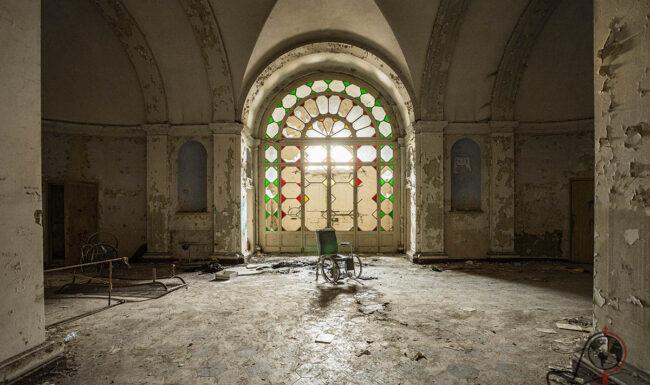 Verlassener Saal im ehemaligen Manicomio di Colorno mit großem farbigem Rundfenster und abblätterndem Wandputz.