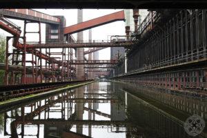Blick auf die Industrieanlagen der Zeche Zollverein in Essen mit roten Förderbrücken, Rohrleitungen und spiegelndem Wasserbecken