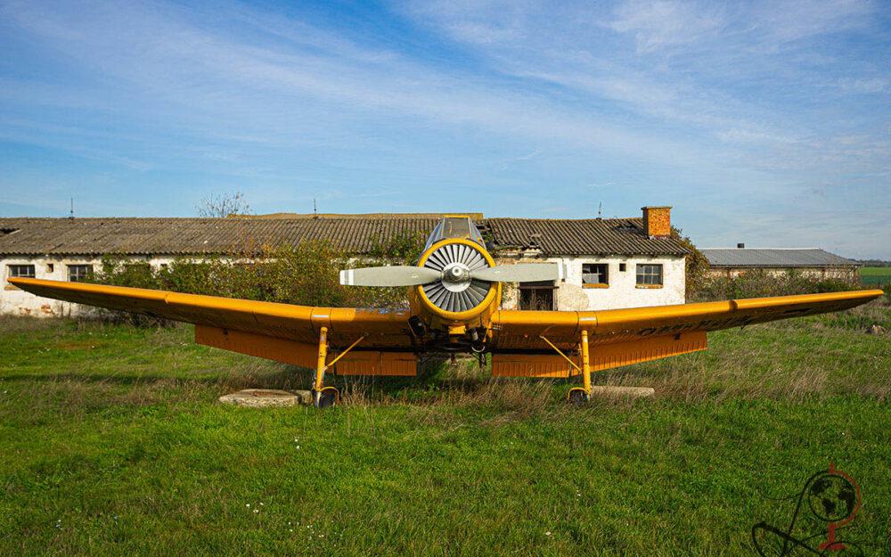 Frontansicht eines gelben Agrarflugzeugs vor verfallenen Hangargebäuden auf einer Wiese