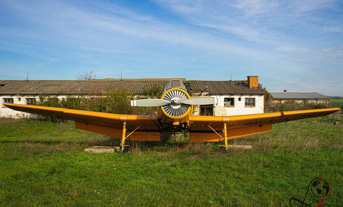 Frontansicht eines gelben Agrarflugzeugs vor verfallenen Hangargebäuden auf einer Wiese