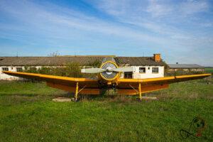 Frontansicht eines gelben Agrarflugzeugs vor verfallenen Hangargebäuden auf einer Wiese