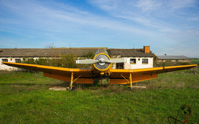 Frontansicht eines gelben Agrarflugzeugs vor verfallenen Hangargebäuden auf einer Wiese